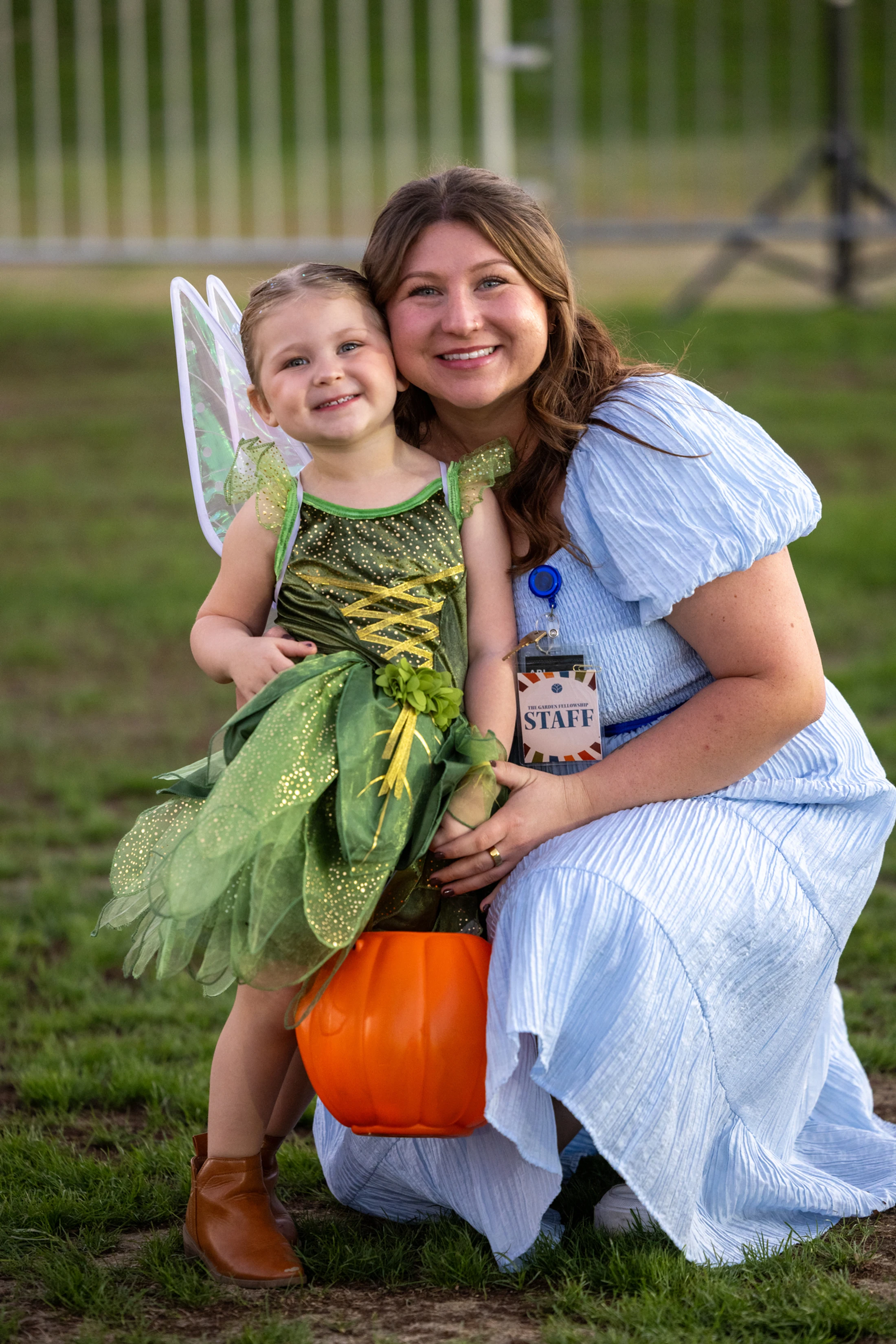 A woman in a blue dress kneels on grass next to a young girl in a green fairy costume holding an orange pumpkin bucket. They are both smiling at the camera.