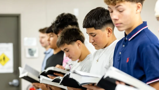 A line of teenage boys stand together in a classroom, each holding an open book and looking down with focused expressions as they read.
