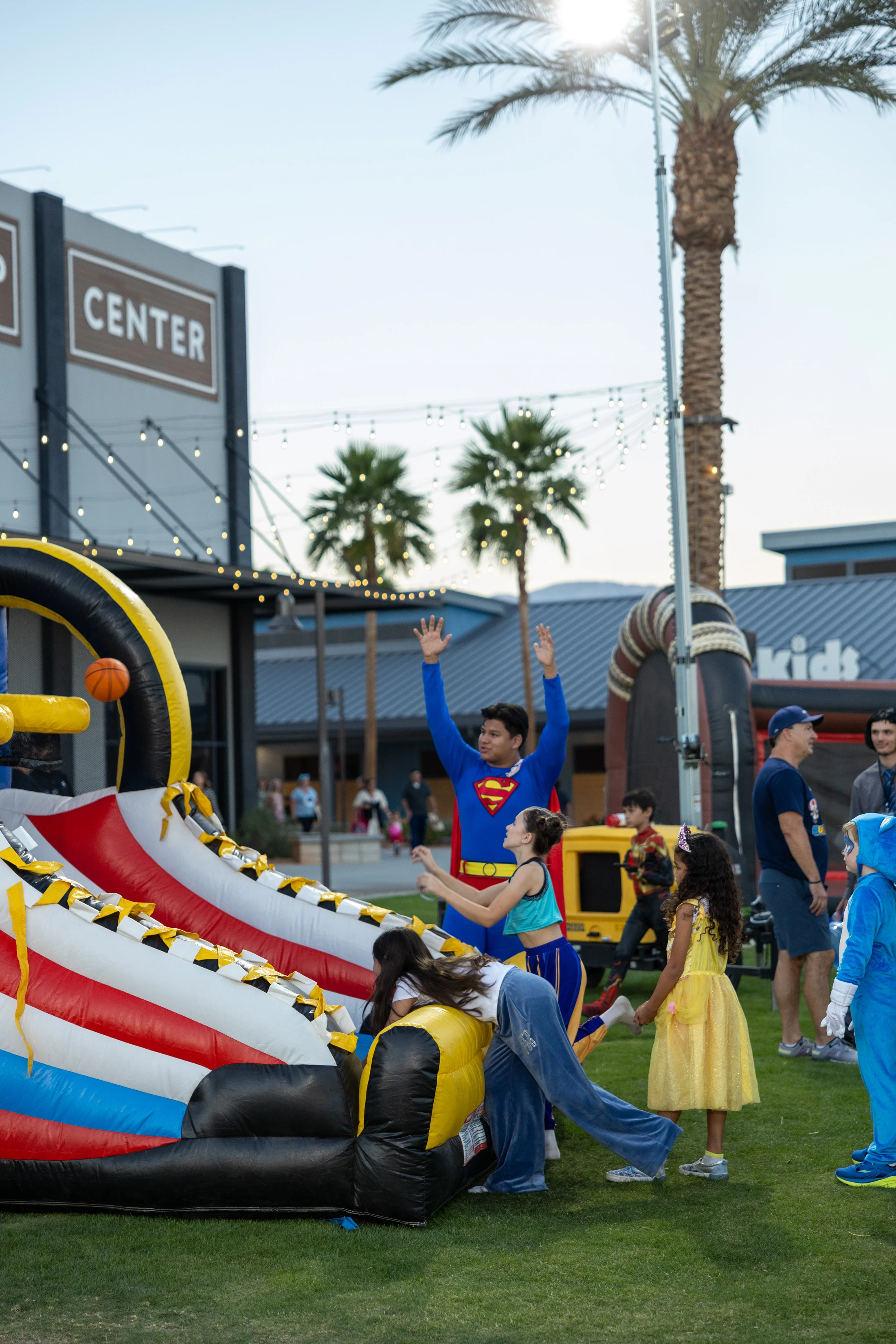 Children and adults in superhero and princess costumes play around a colorful inflatable game at an outdoor festival with palm trees in the background.