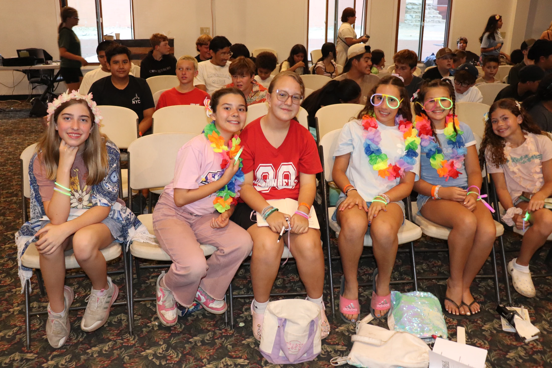 A group of six young girls sit in the front row at an event, smiling for the camera. Some wear colorful leis, glow glasses, and a flower crown.
