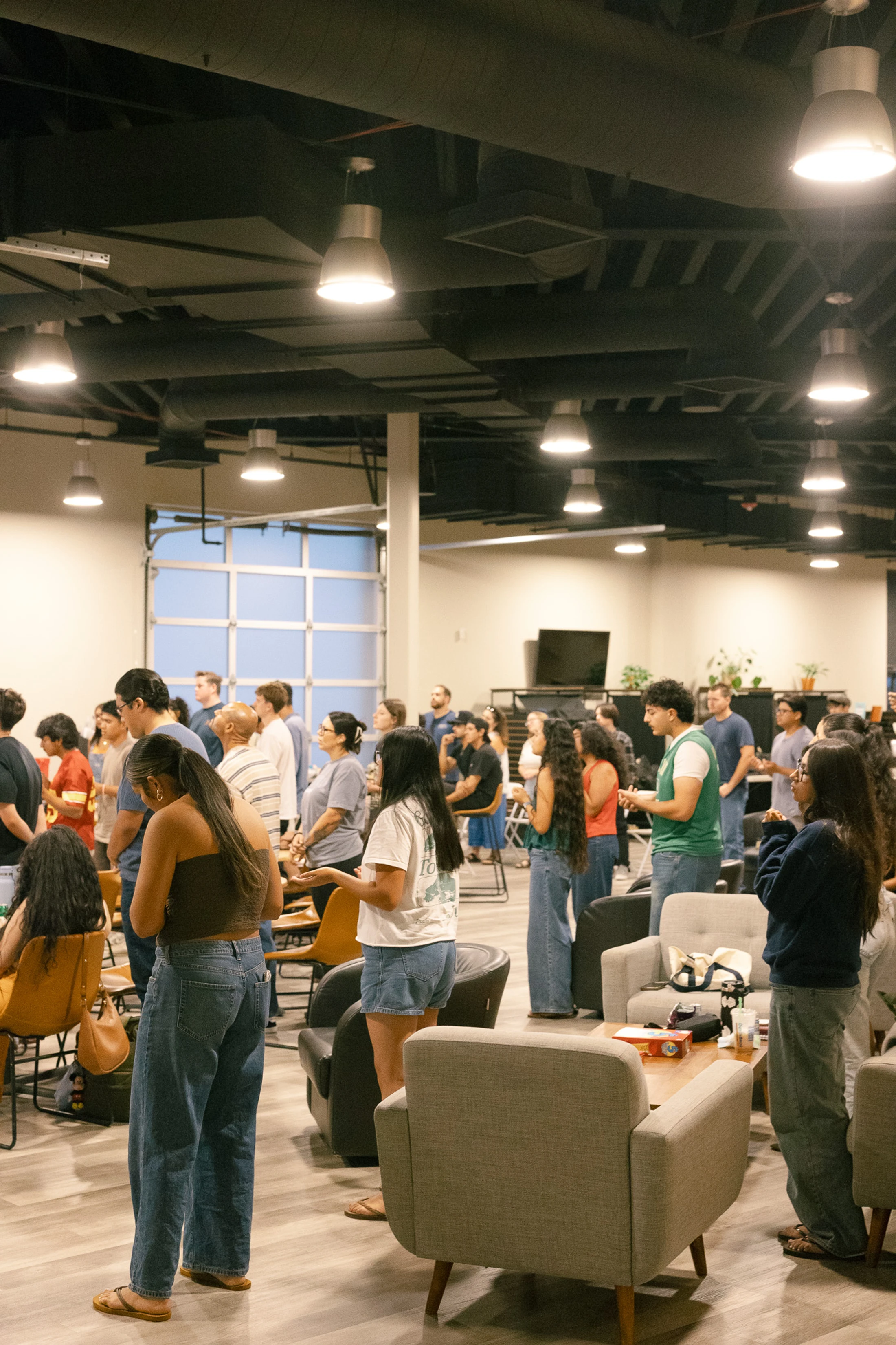 A diverse group of casually dressed people stand in a large, modern room with industrial lighting, attentively facing forward during a gathering.