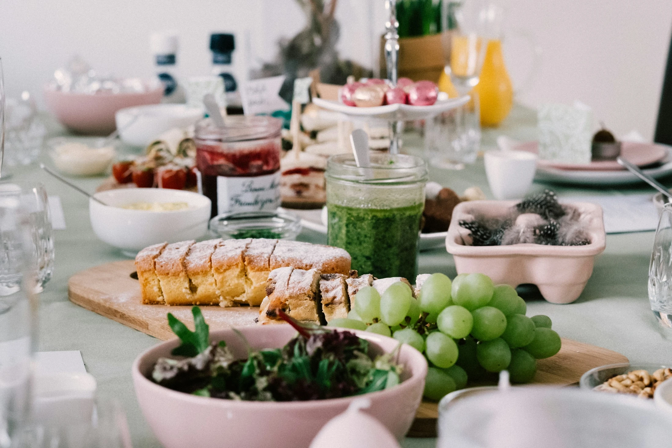 A beautifully set table for brunch, featuring a sliced loaf cake on a wooden board, fresh grapes, salad, pesto, jam, and other festive dishes.