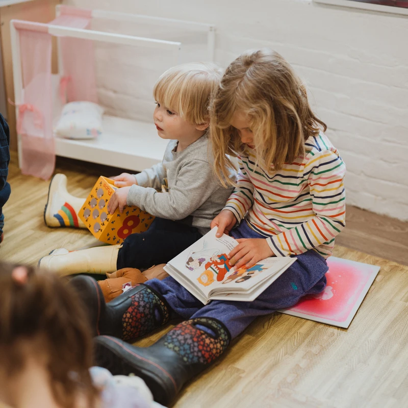 A young girl and a younger boy sit side-by-side on a wooden floor, looking at picture books together in a classroom setting.