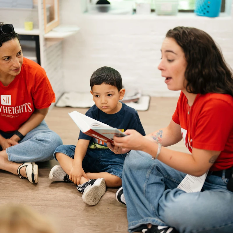 Two women sit on the floor with a young boy between them. One woman reads from a book while the boy listens intently, and the other woman watches them.
