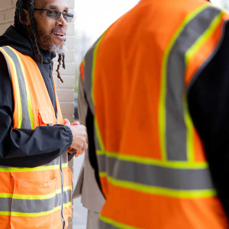 A man with dreadlocks and glasses wearing an orange safety vest smiles while talking to another person whose back is to the camera, also in a safety vest.