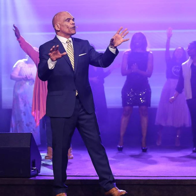 A bald man in a navy suit and striped tie sings on stage, gesturing with his hands. Behind him, a choir sings under purple and white lights.