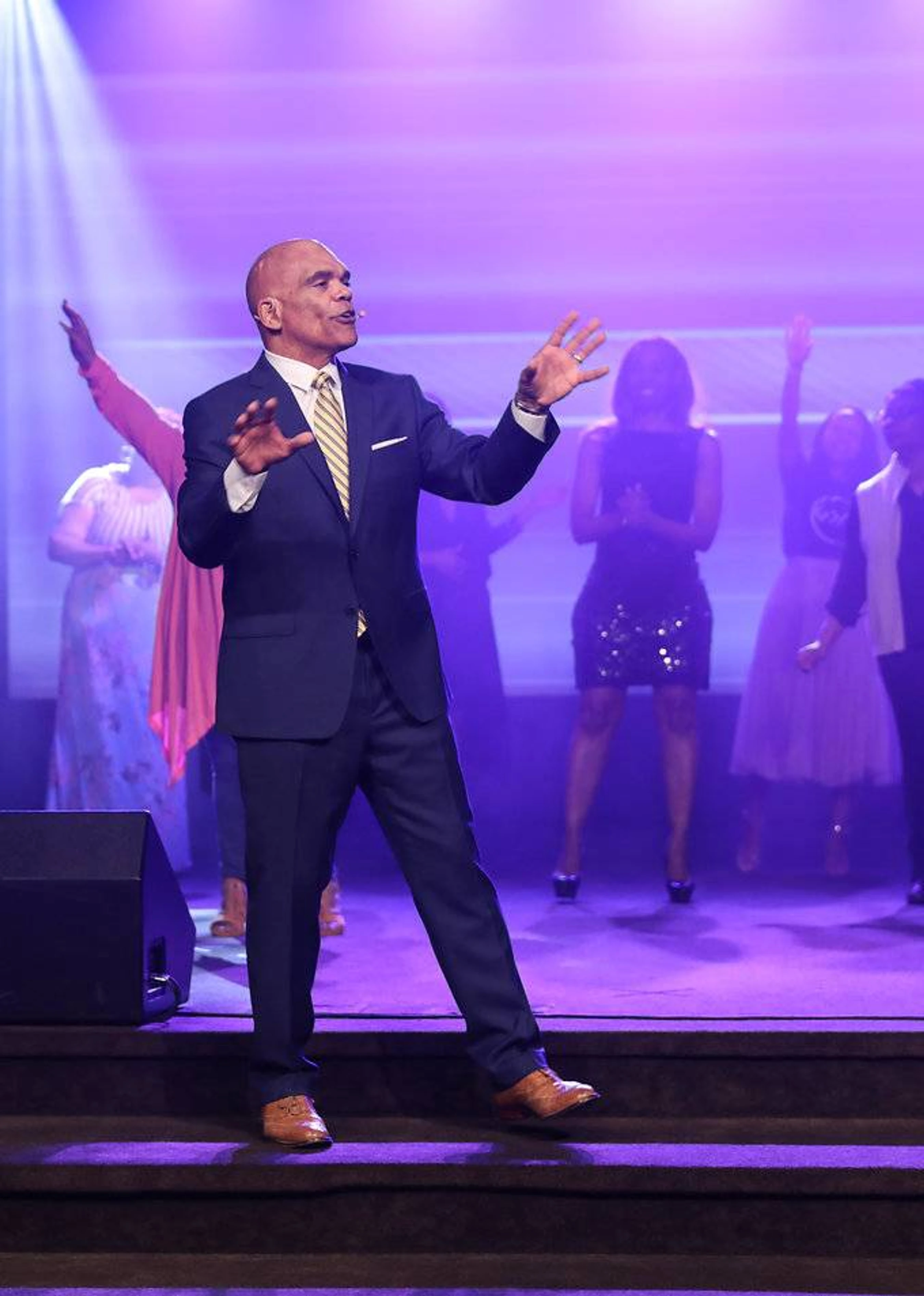 A bald man in a navy suit and striped tie sings on stage, gesturing with his hands. Behind him, a choir sings under purple and white lights.