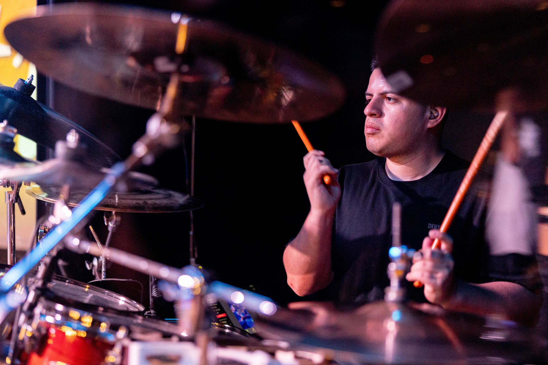 A male drummer in a black t-shirt looks focused as he plays his drum kit on a dimly lit stage, holding bright orange drumsticks in mid-air.