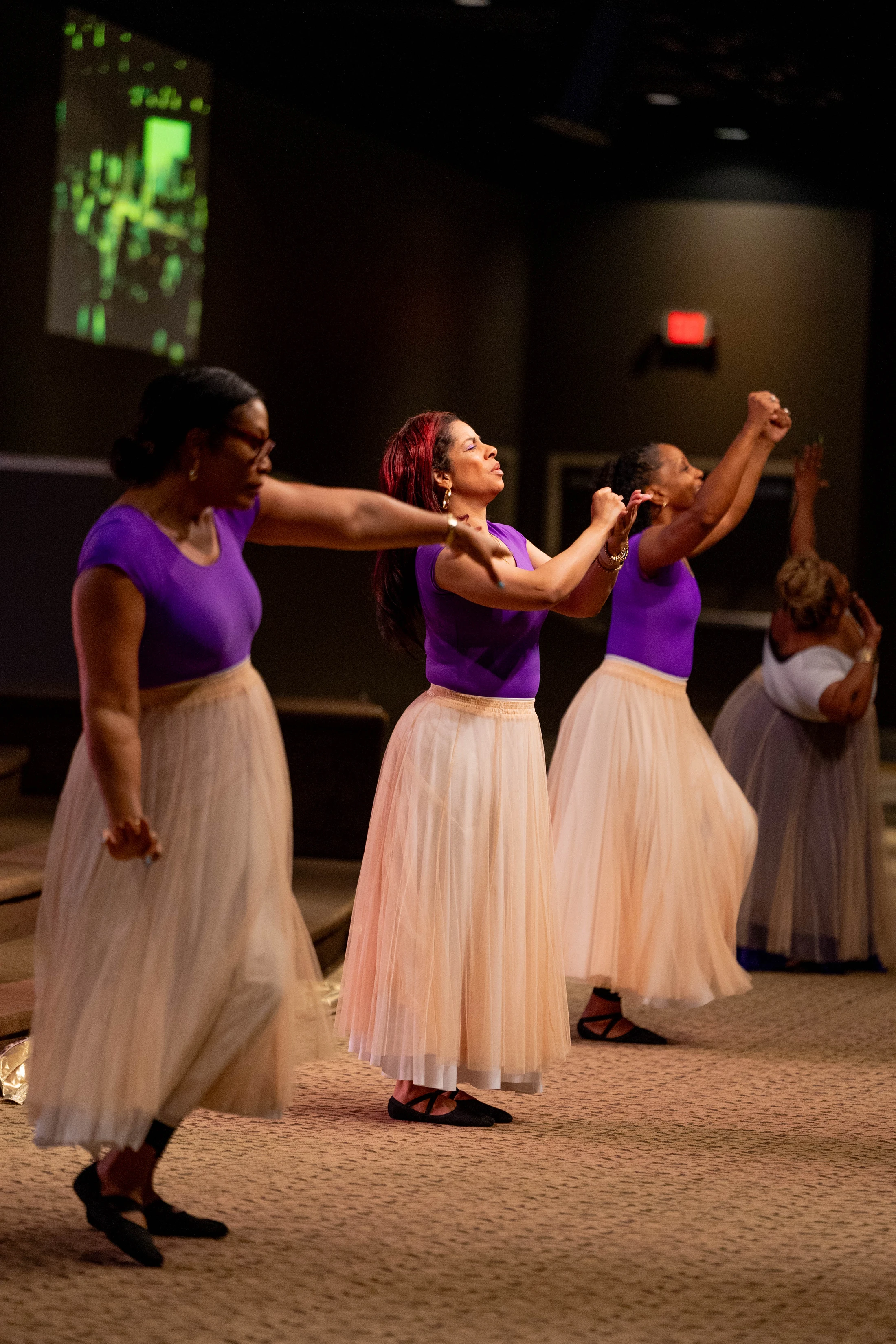 A line of women in matching purple tops and long, cream-colored tulle skirts perform an expressive dance on a carpeted stage in a dimly lit room.