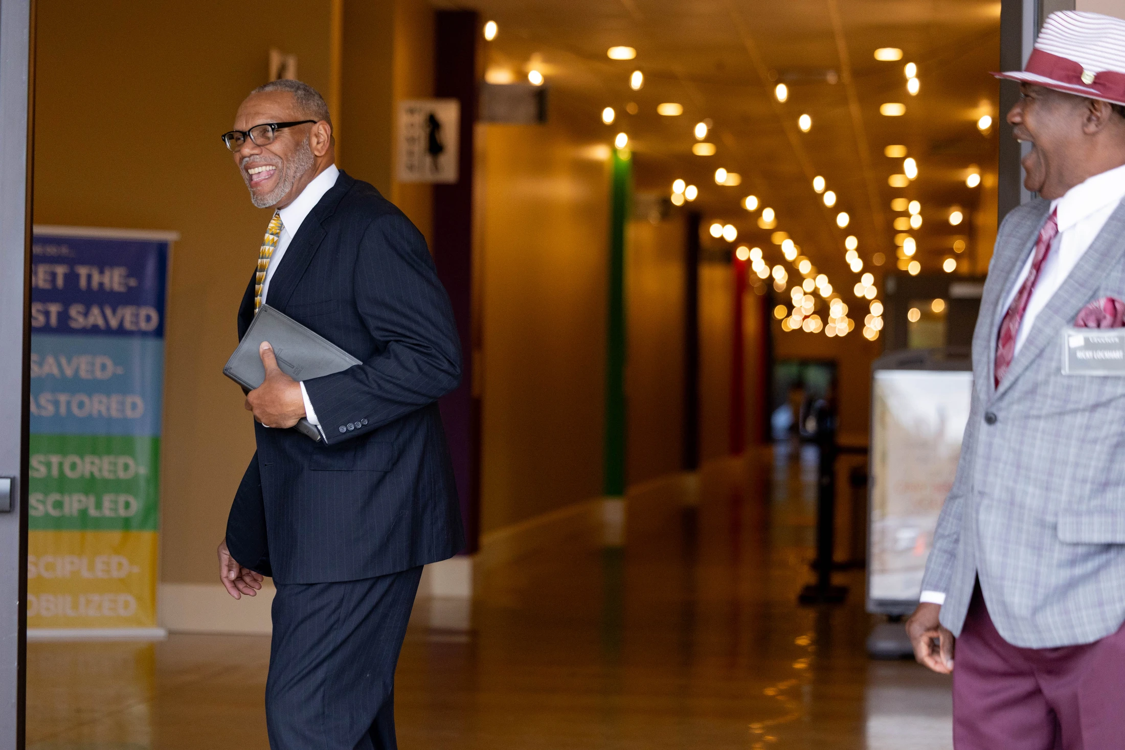 A man in a pinstripe suit and glasses smiles as he walks through a doorway, holding a bible. Another man in a hat and blazer smiles beside him in a hallway.