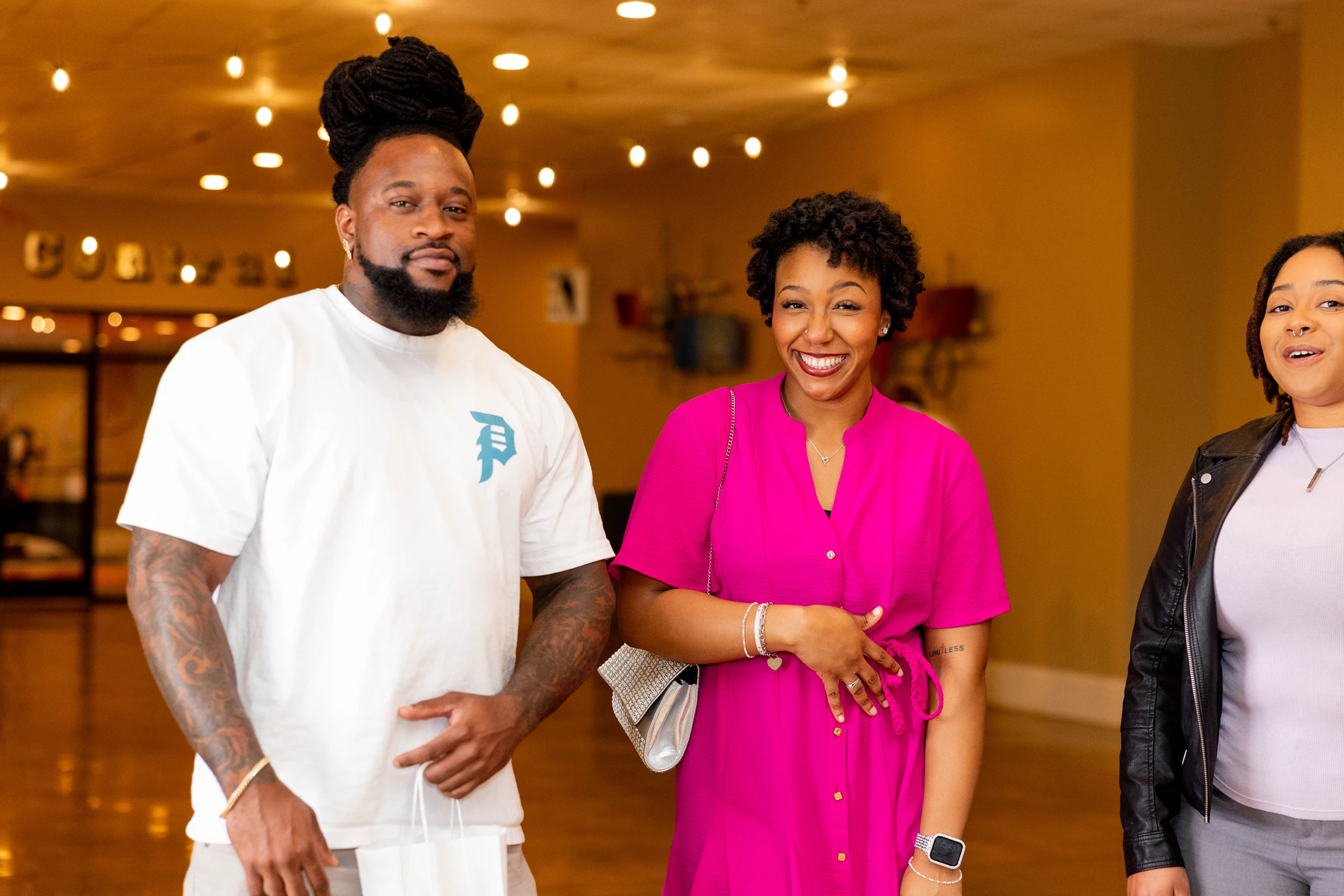 A Black man with tattoos and two Black women stand together indoors, smiling. The woman in the middle wears a bright pink dress. The background is warmly lit.