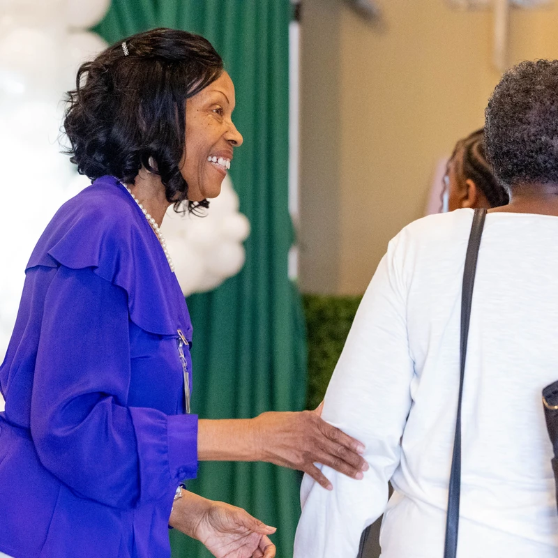 A smiling woman in a bright blue blouse and pearl necklace reaches out to greet another woman, who is seen from behind, during an indoor event.