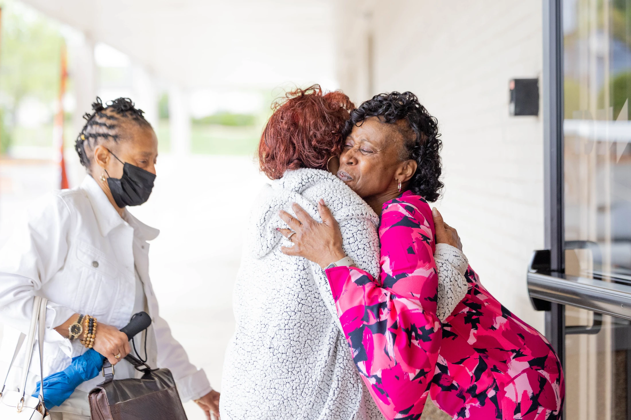 Two older Black women share a warm hug while a third woman in a face mask looks on. They are standing outdoors under a covered walkway.