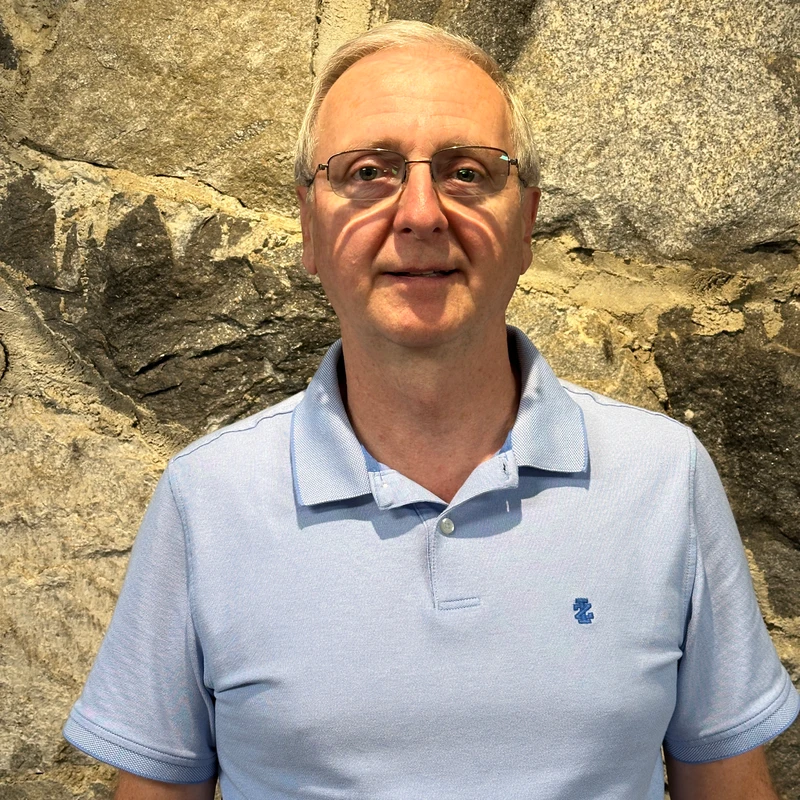 A middle-aged man with short, light hair and glasses wearing a light blue polo shirt stands in front of a rough, sunlit stone wall, looking at the camera.