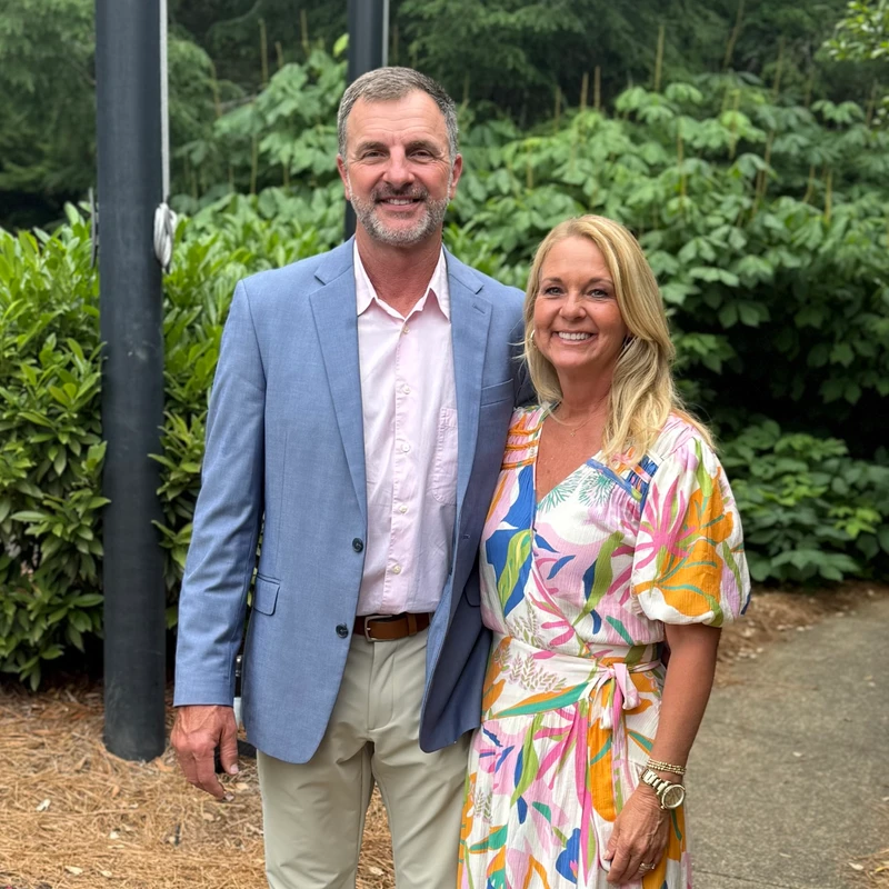 A man in a blue blazer and a woman in a colorful patterned dress stand together smiling for a photo in front of a background of green bushes.