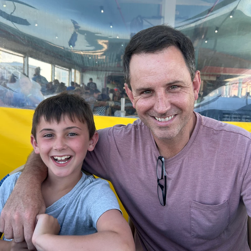 A man with dark hair and a pink shirt has his arm around a smiling young boy in a grey shirt. They are sitting together at a red table, looking at the camera.