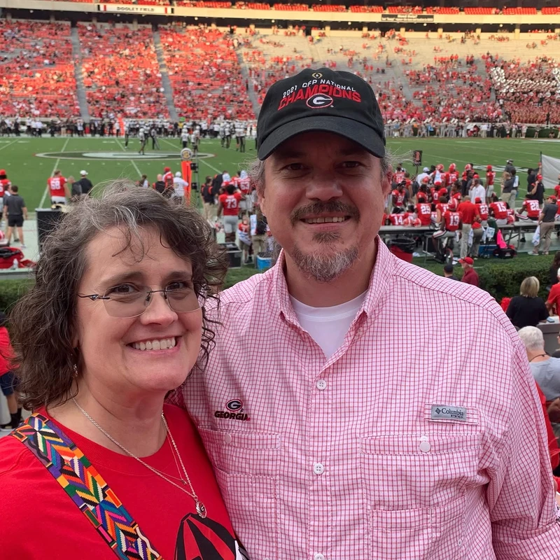 A smiling man and woman pose for a photo in the stands of a crowded football stadium, both wearing Georgia Bulldogs fan gear.