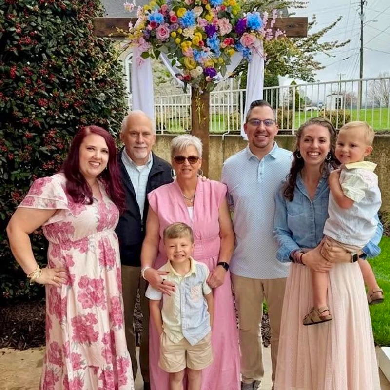 A family of seven, spanning three generations, smiles for a photo outdoors in front of a wooden cross adorned with colorful flowers for Easter.
