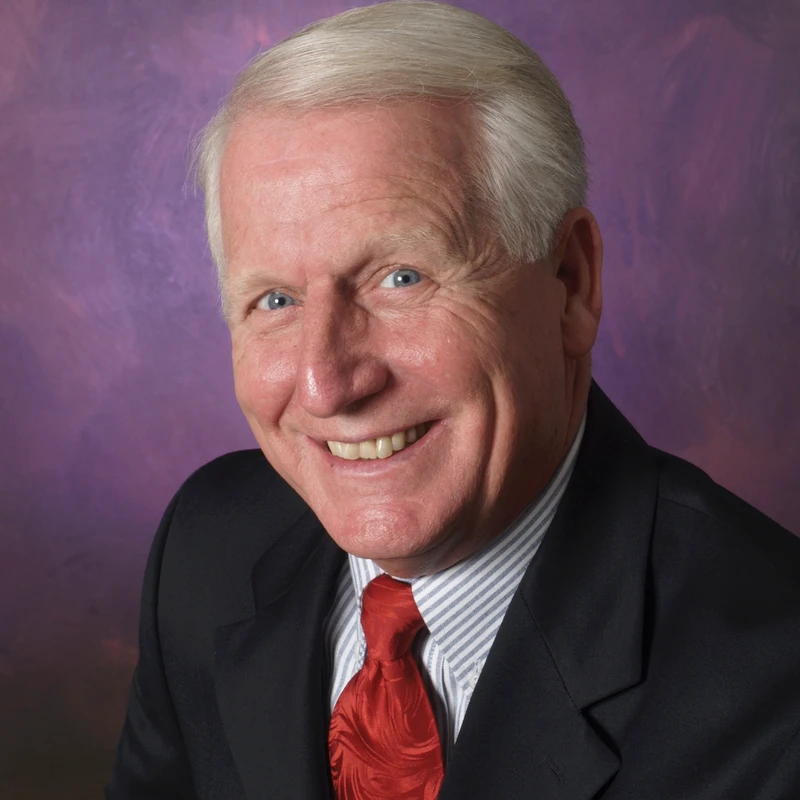 A head-and-shoulders portrait of a smiling older man with white hair and blue eyes, wearing a suit and red tie against a purple studio background.