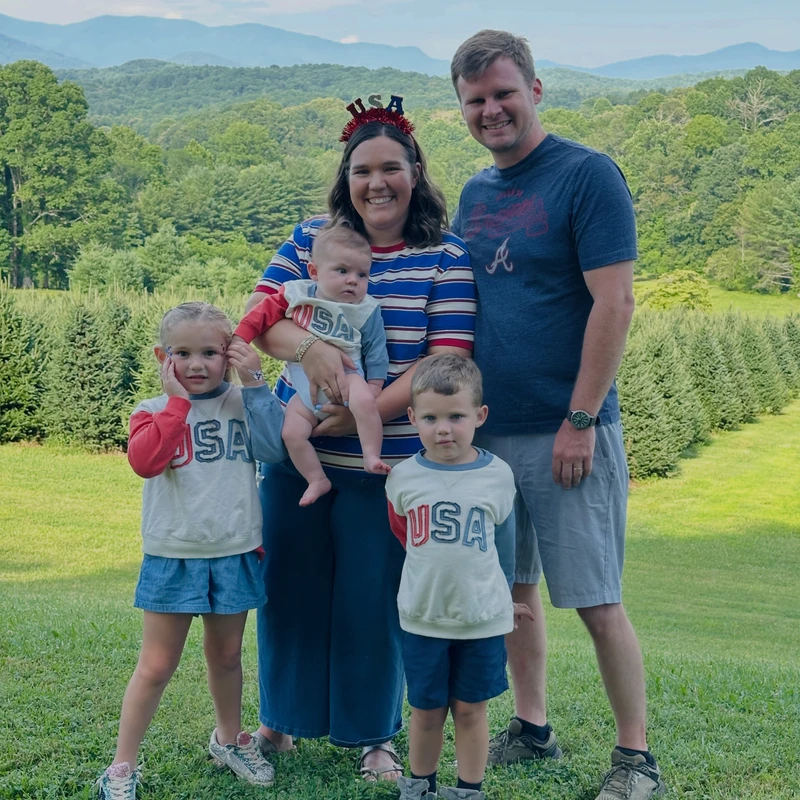 A smiling family of five in patriotic 'USA' outfits stands in a grassy field for a portrait, with a backdrop of rolling green hills and distant blue mountains.