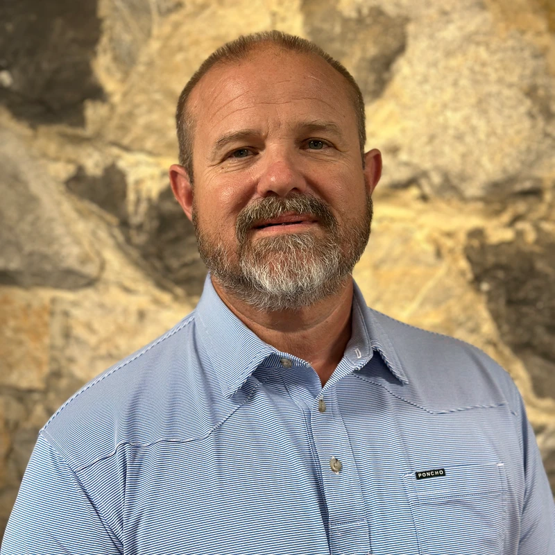 A head-and-shoulders portrait of a middle-aged man with a beard, wearing a blue and white striped polo shirt and standing in front of a stone wall.