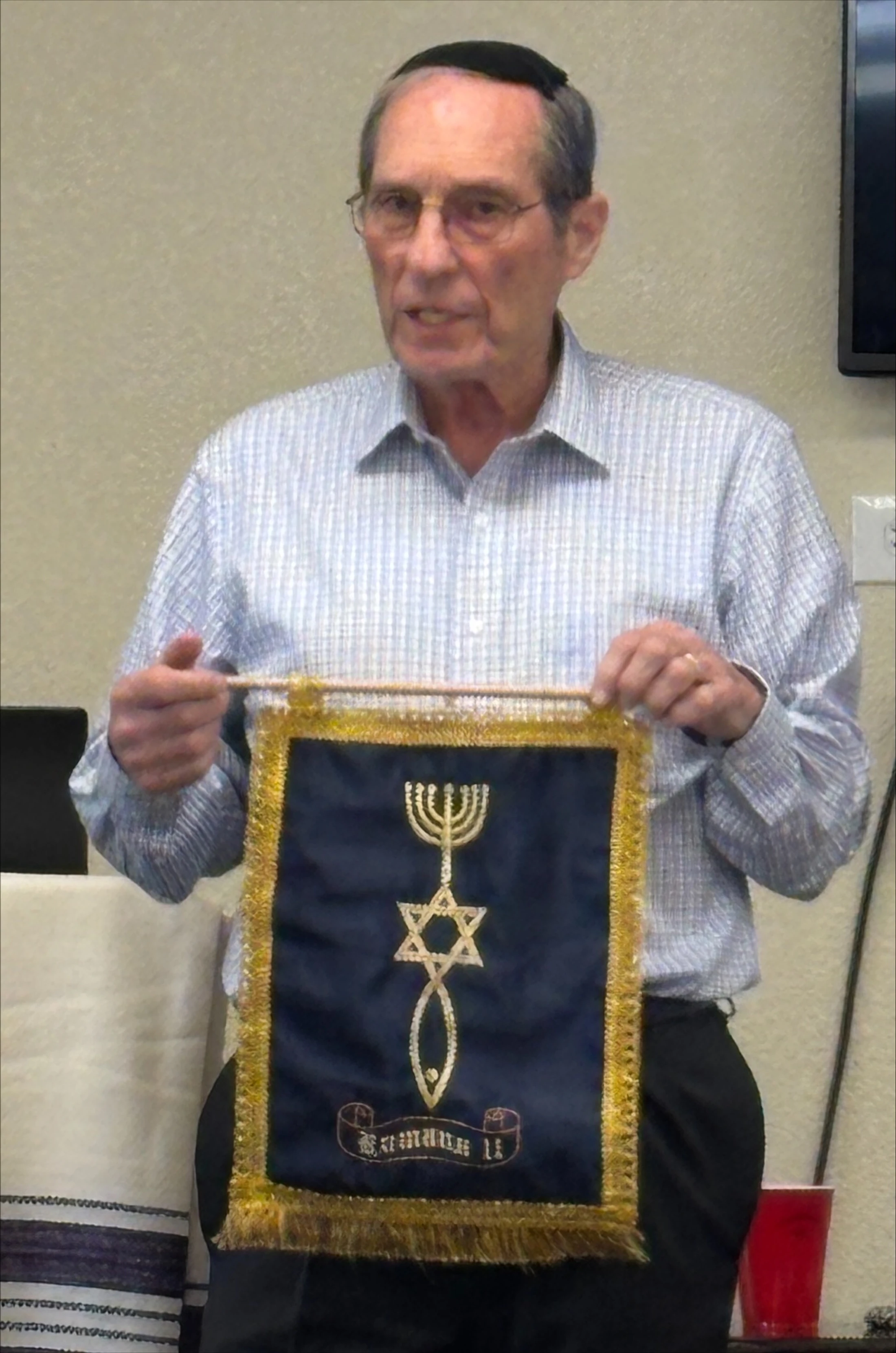 An older man wearing a yarmulke holds up a small blue banner with gold fringe. It has a menorah, Star of David, and a Christian fish symbol embroidered on it.