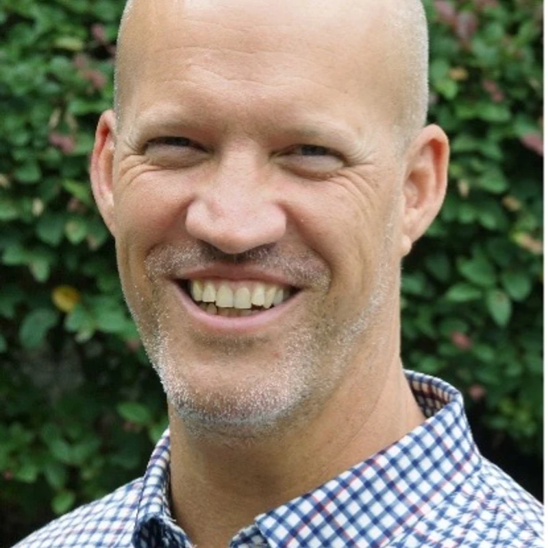A head and shoulders portrait of a smiling bald man with light stubble. He wears a blue and white checkered shirt and stands in front of green foliage.