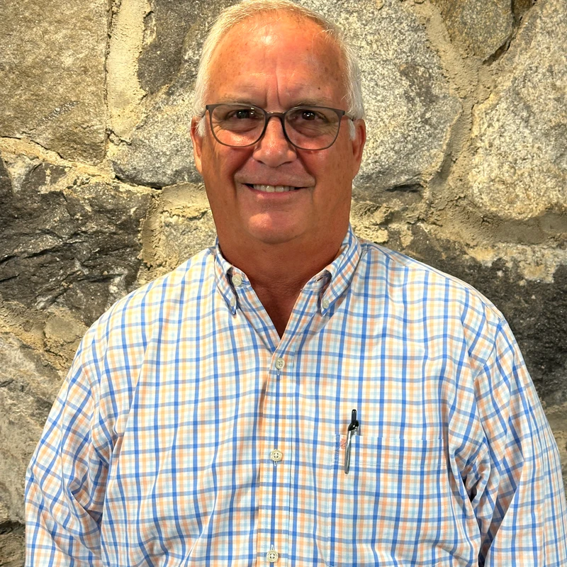 A head-and-shoulders portrait of a smiling older man with white hair and glasses, wearing a blue and orange plaid shirt, standing before a stone wall.