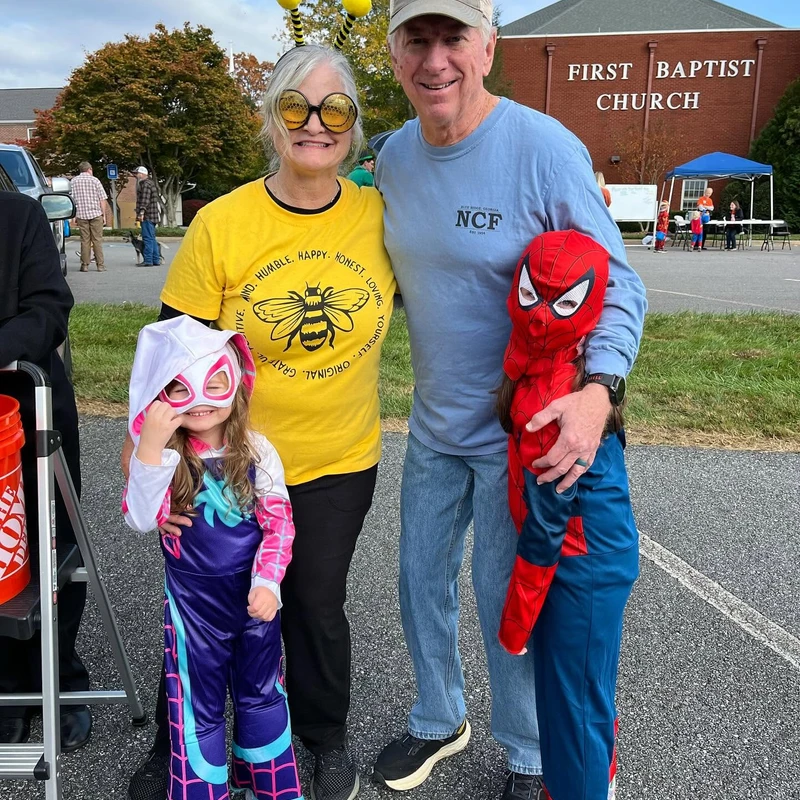 A couple poses with two small children dressed in superhero costumes in a church parking lot. The woman wears a bee costume and the kids are Spider-Man.