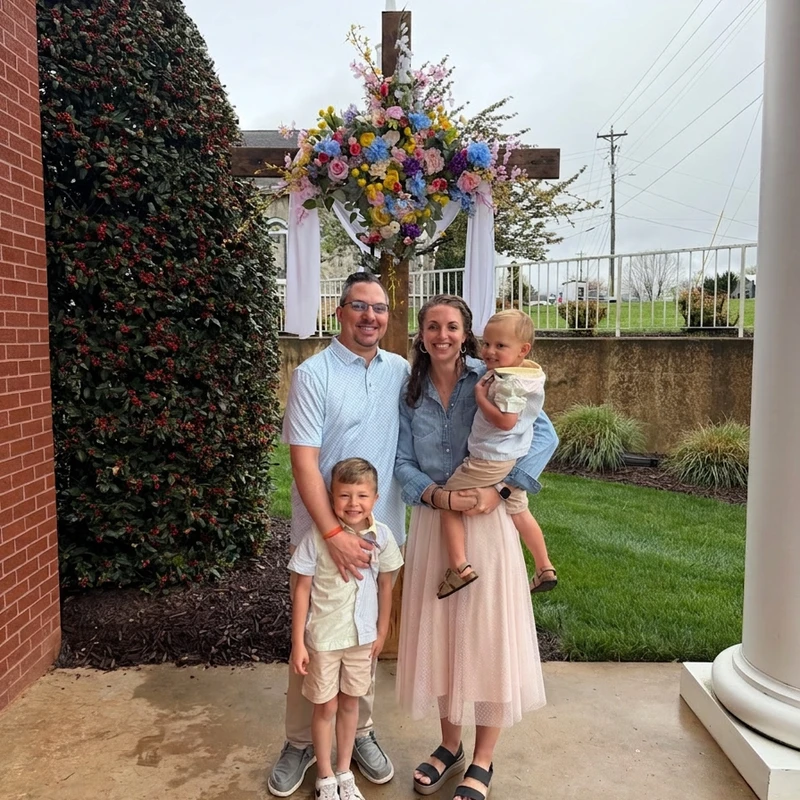 A smiling family of four, a man, a woman, and two young boys, stand for a photo in front of a large wooden cross adorned with colorful flowers.