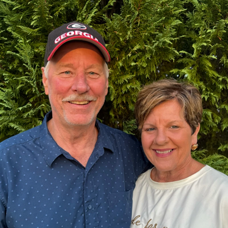 A smiling man and woman stand close together in front of a green hedge. The man wears a blue shirt and a Georgia baseball cap, with his arm around the woman.