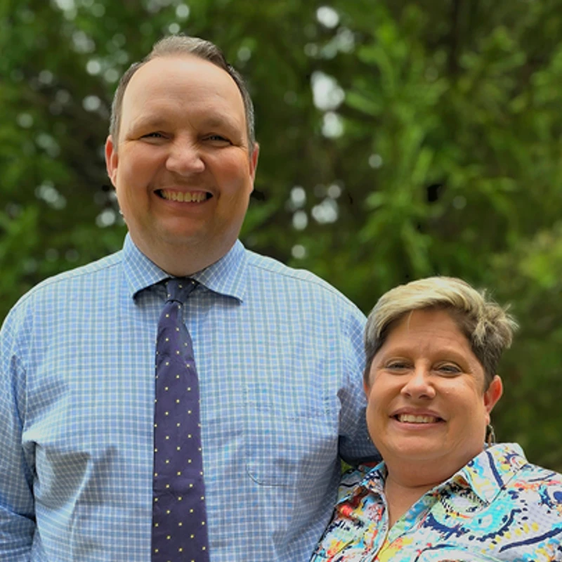 A man in a blue checked shirt and tie stands smiling next to a woman with short hair and a colorful shirt, posing in front of green trees.