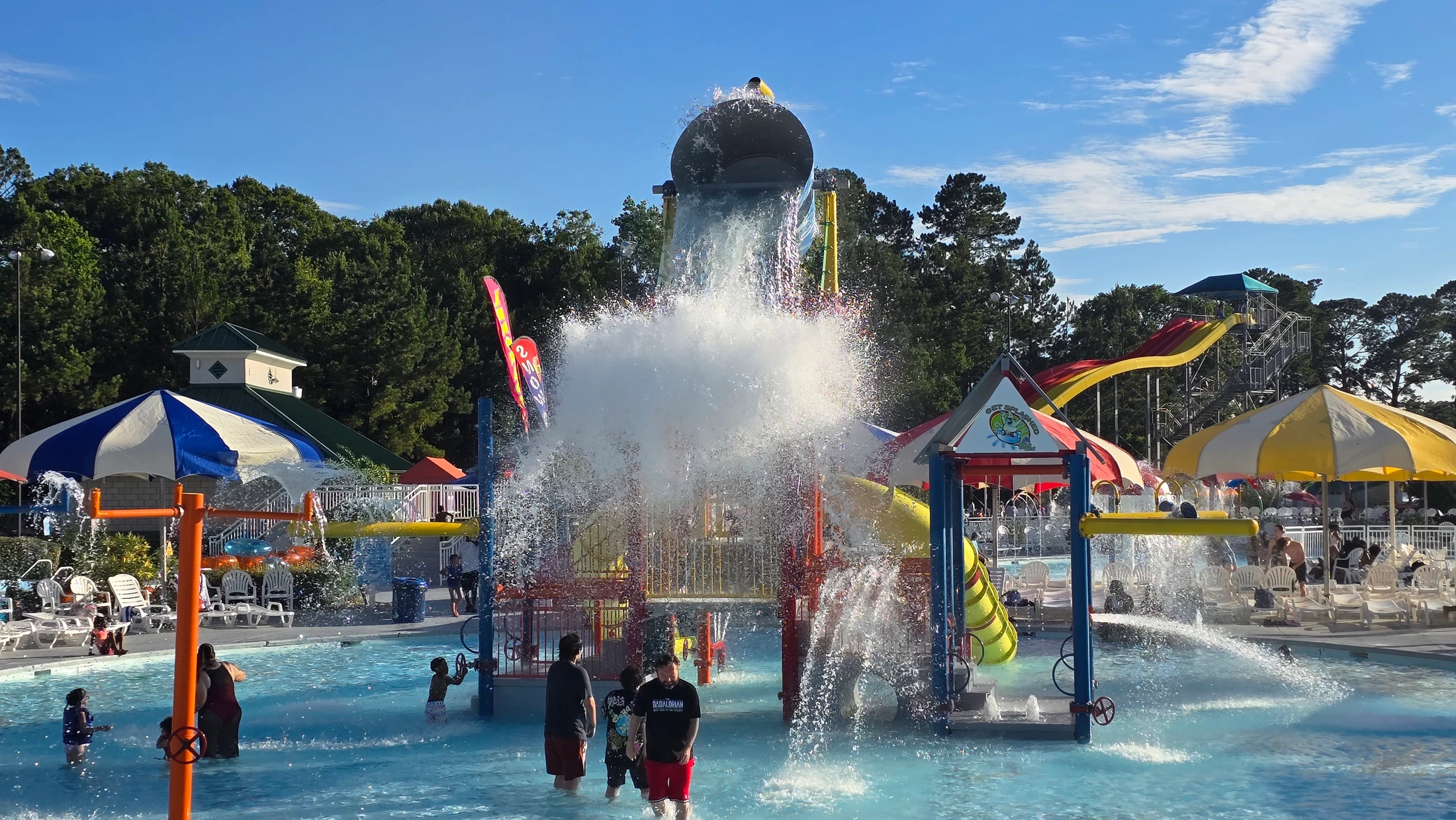 A large tipping bucket pours water onto a colorful play structure, creating a huge splash for families playing in the pool below on a sunny day.