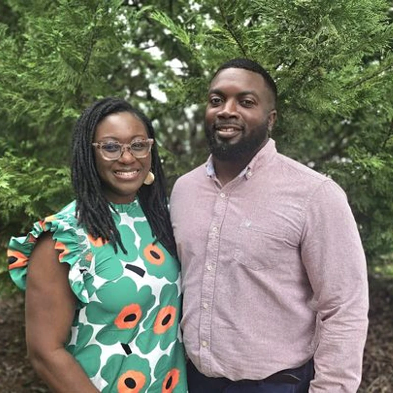 A Black man and woman smiling at the camera while standing in front of a large evergreen tree. She wears a floral dress and he wears a pink button-down shirt.