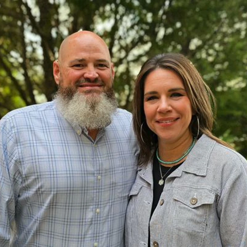 A bald man with a grey beard and a woman with brown hair smile for a photo outdoors. The man wears a plaid shirt and the woman wears a striped jacket.