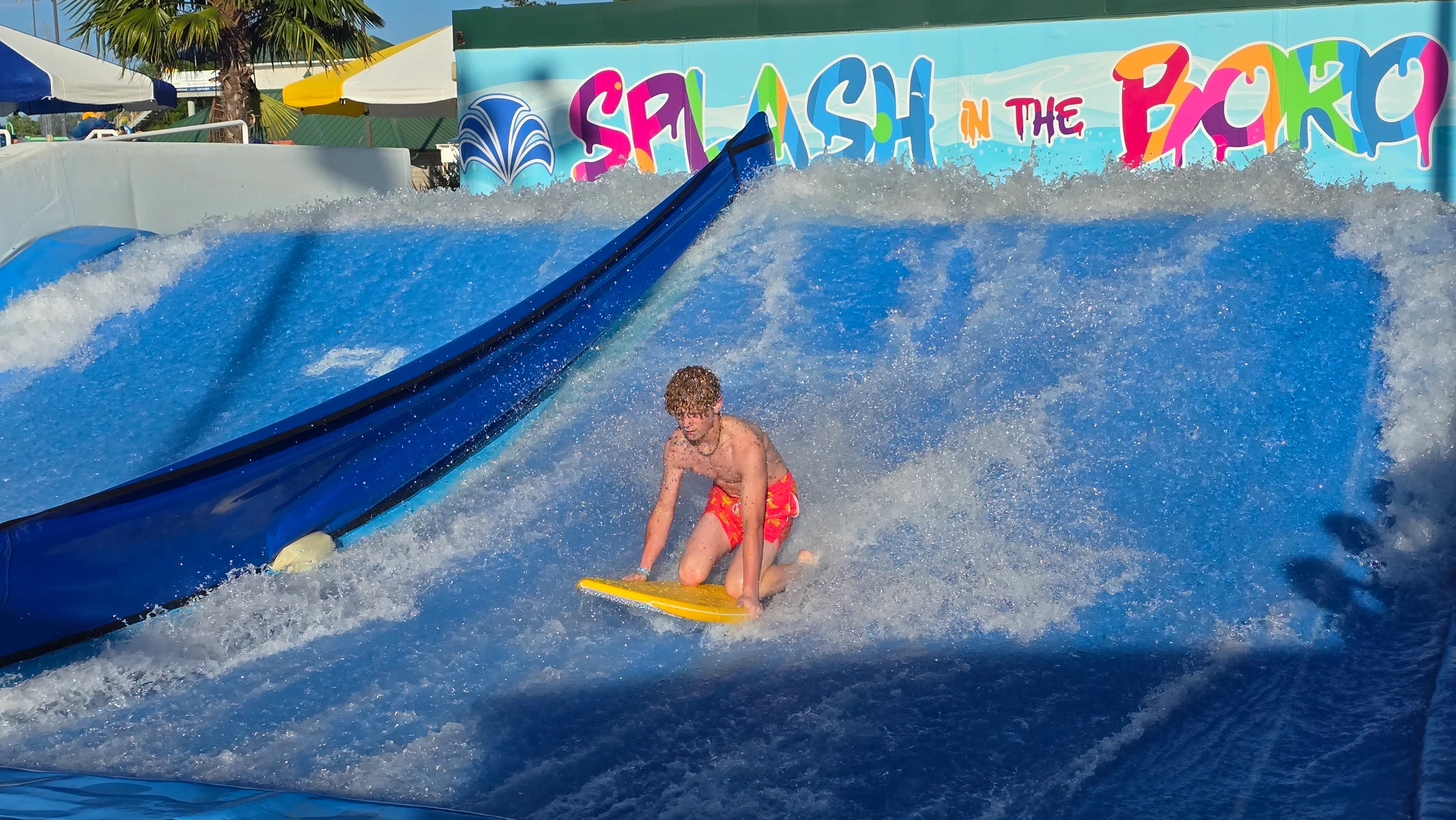 A young man with curly hair kneels on a yellow bodyboard, riding a wave on a surf simulator at the Splash in the Boro water park.