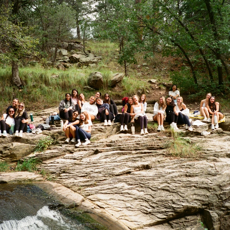 A large group of young women are sitting together on a layered rock formation next to a small waterfall, smiling for a photo in a dense forest.