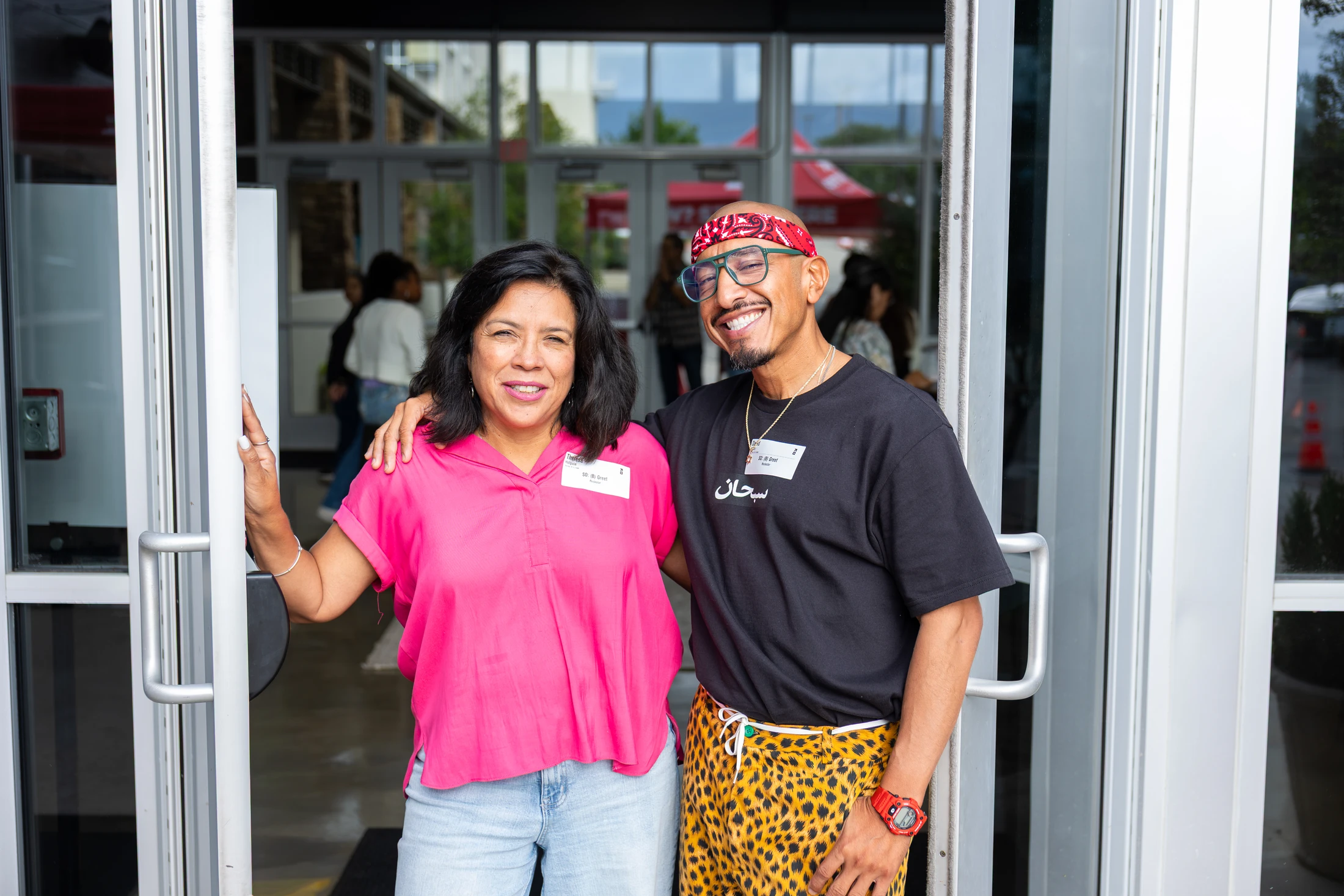 A smiling woman in a pink shirt and a man in a black tee and cheetah-print pants pose with their arms around each other in an open glass doorway.