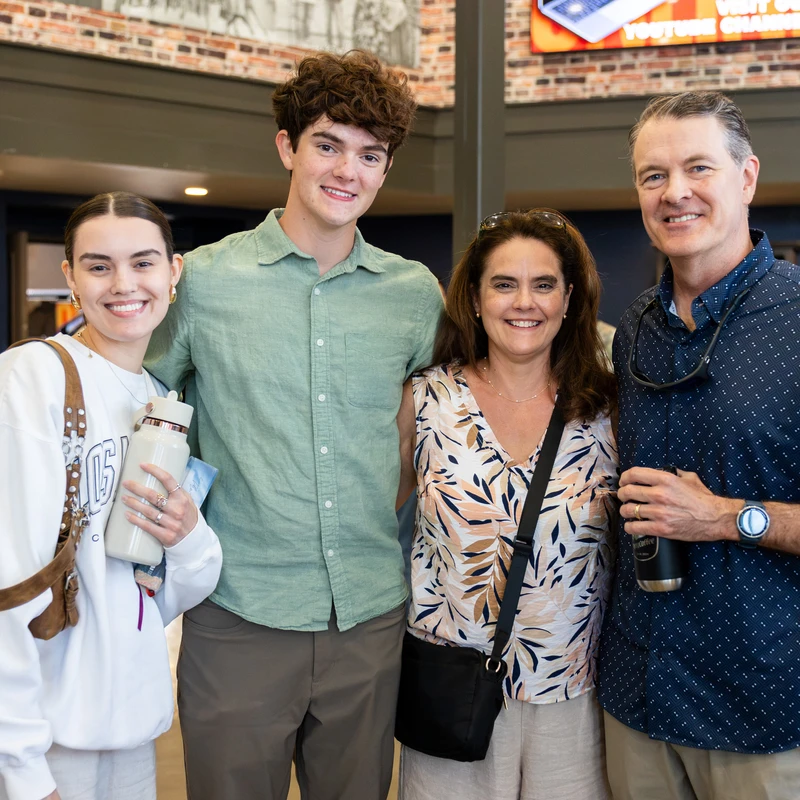 A family of four, parents and two teenage children, stand side-by-side smiling at the camera while posing for a picture in a building lobby.