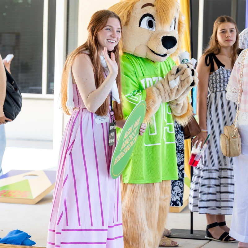 A smiling woman in a pink and white striped dress waves while posing next to a furry mascot in a bright green jersey at an outdoor event.