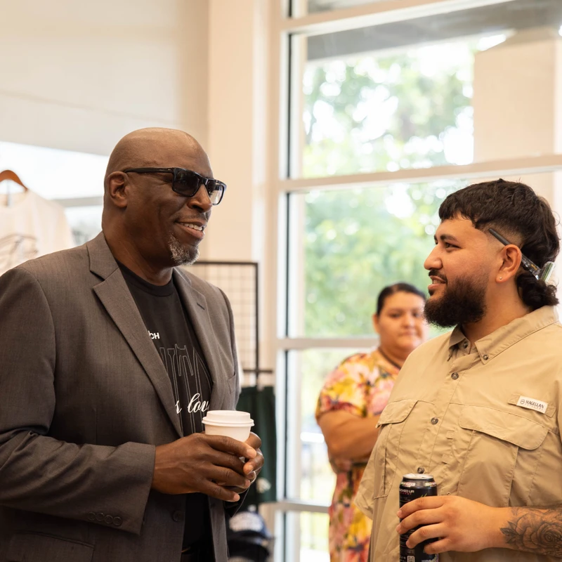 An older Black man in a blazer and a younger man with a mullet haircut smile as they talk. The older man holds a coffee cup and the younger holds a drink can.
