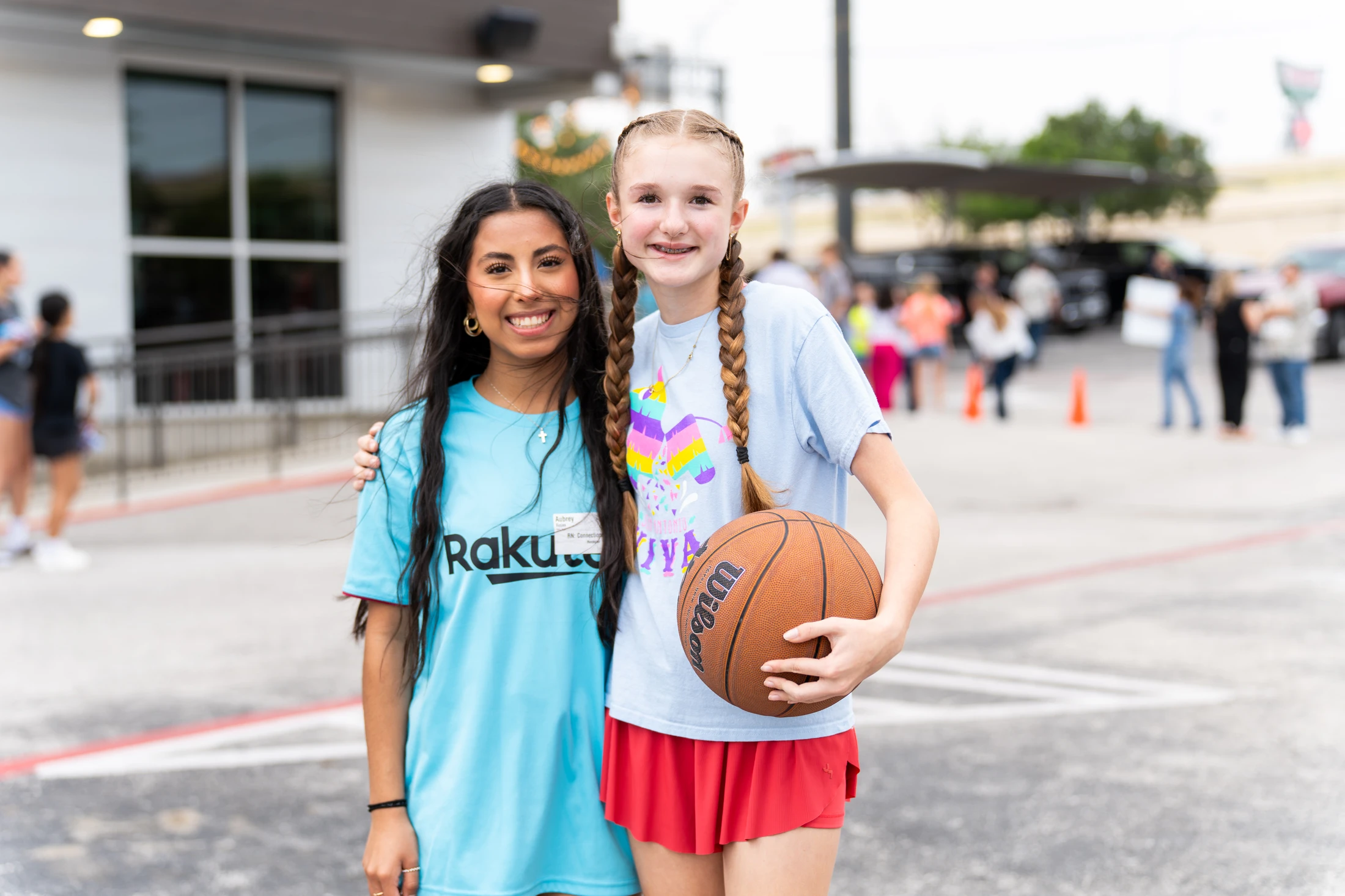 A young woman in a blue Rakuten volunteer t-shirt smiles with her arm around a girl with pigtail braids who is holding a basketball outdoors.