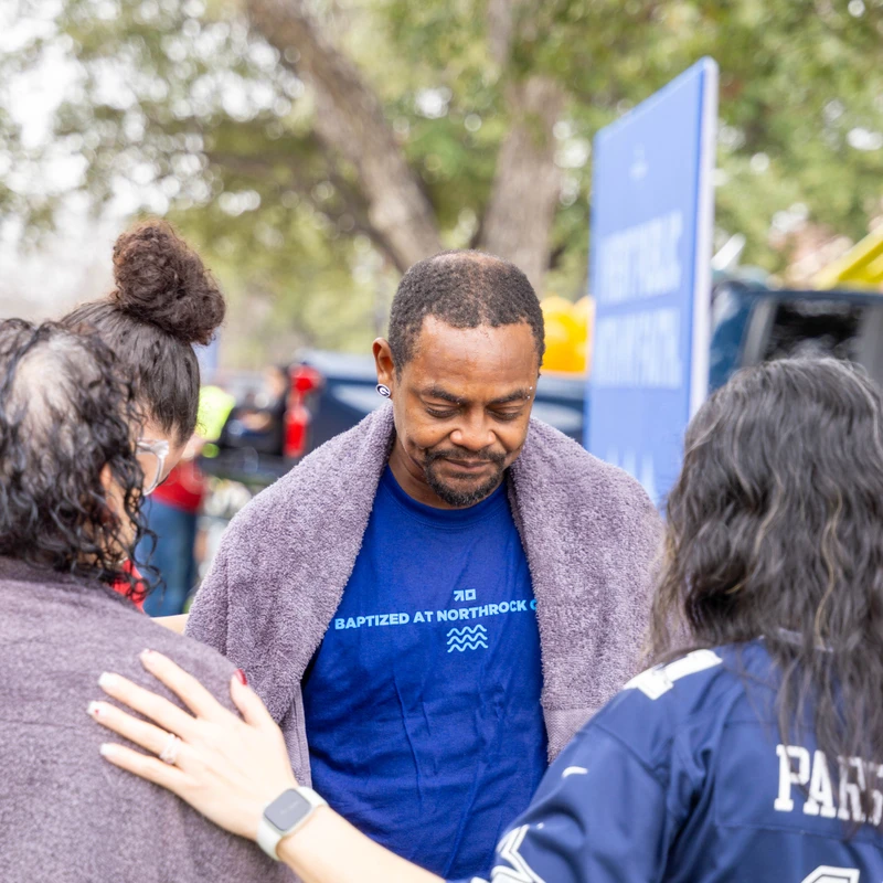 A man in a blue 'Baptized' t-shirt looks down with a towel on his shoulders while two women, with their backs to the camera, pray with him outdoors.
