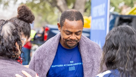 A man in a blue 'Baptized' t-shirt looks down with a towel on his shoulders while two women, with their backs to the camera, pray with him outdoors.