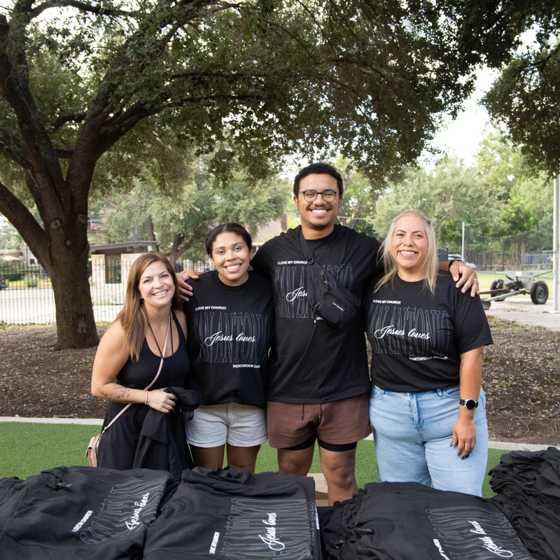 A group of four volunteers, three women and one man, smile while standing behind a table of black t-shirts with 'Jesus loves' written on them.