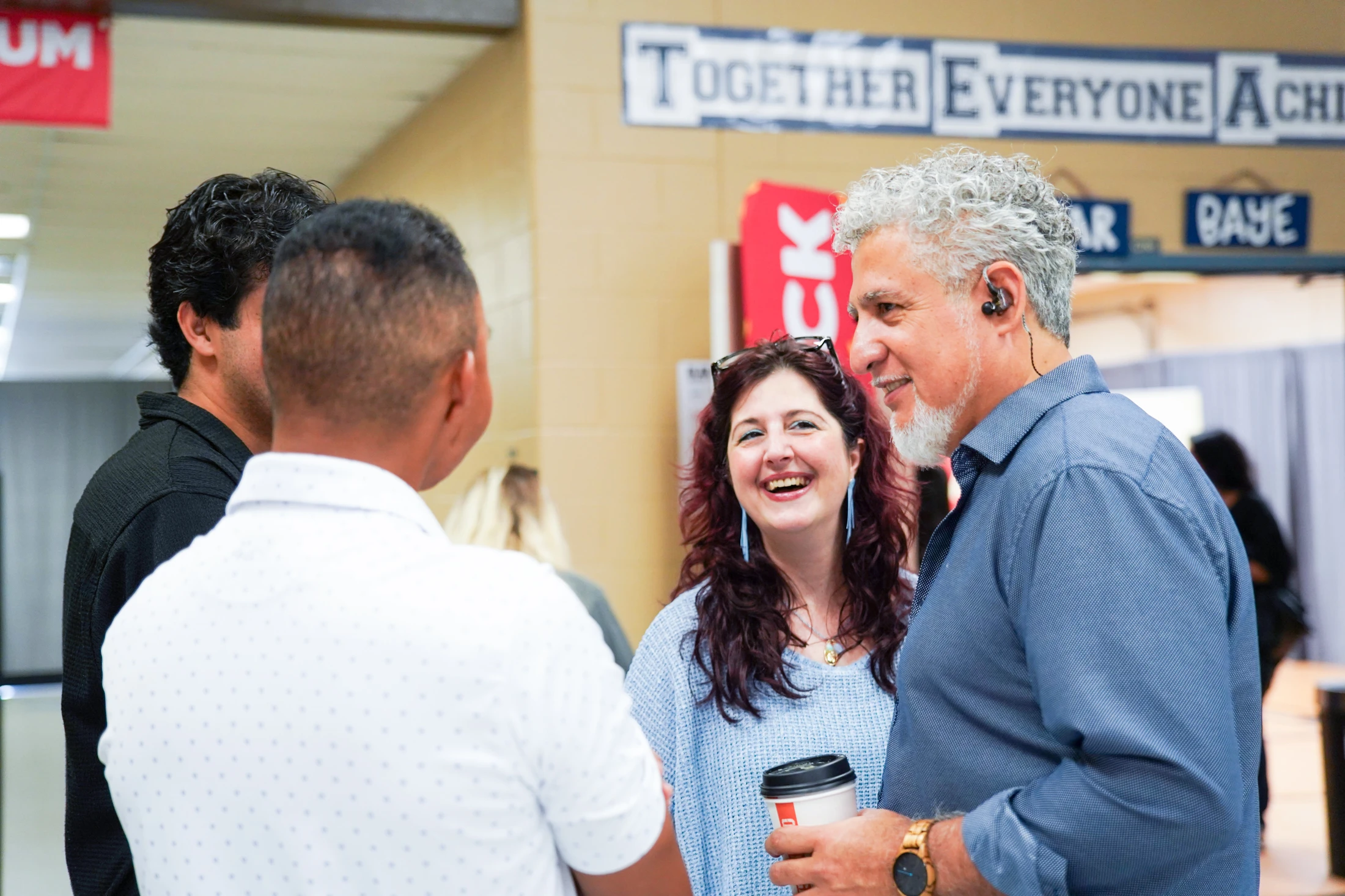 A diverse group of colleagues talking in an indoor space. A woman in the center is smiling broadly while a man with grey hair listens and holds a coffee cup.