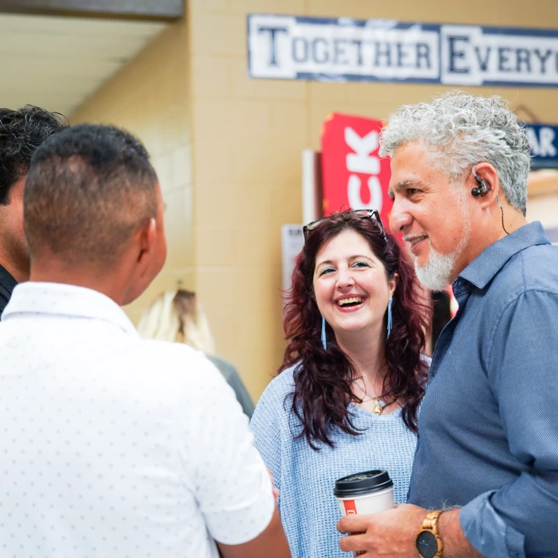 A diverse group of colleagues talking in an indoor space. A woman in the center is smiling broadly while a man with grey hair listens and holds a coffee cup.