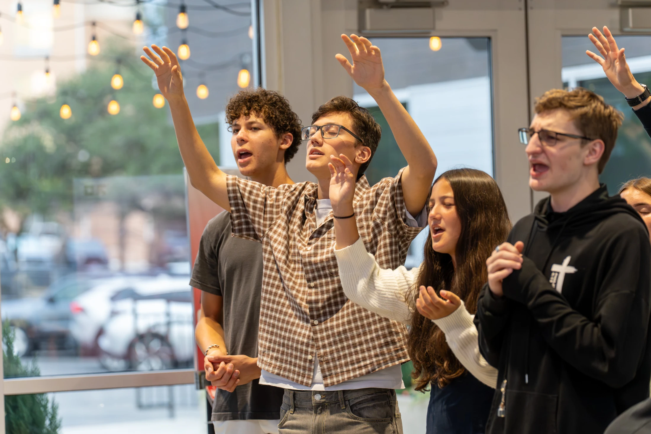 A diverse group of teenagers stand indoors, singing with their hands raised in an expression of worship in front of a large window.