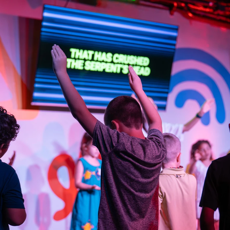 A young boy, seen from behind, raises his hands in worship at a children's event. Other kids are around him, facing a stage with song lyrics on a screen.