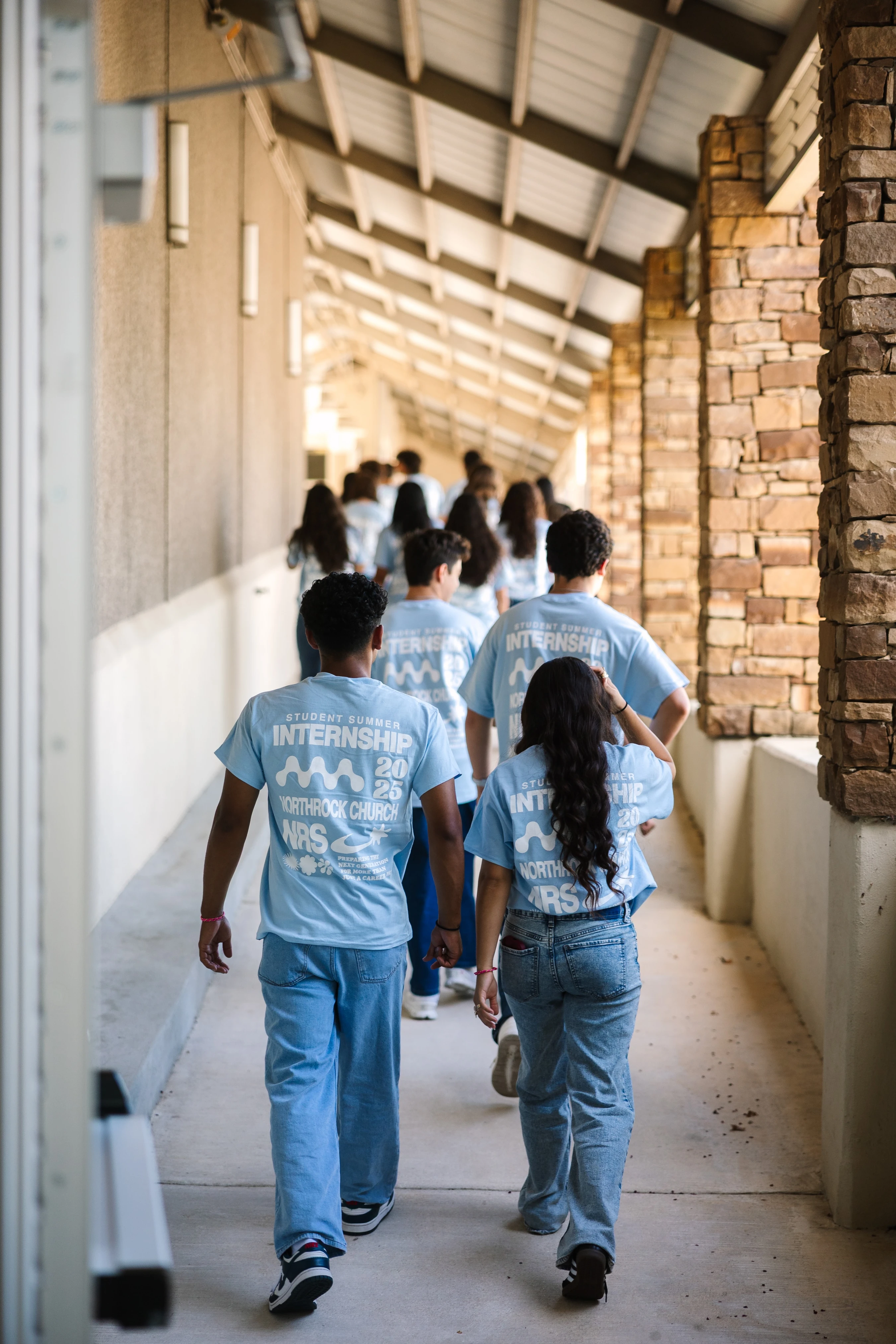 A group of young people, seen from behind, walk down a covered outdoor corridor wearing matching light blue t-shirts for a student summer internship.